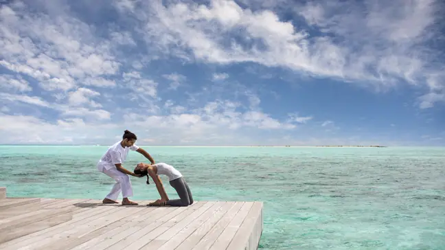 A woman on her knees, bending over backward, with help from a masseuse. On a pavilion overlooking the ocean in the Maldives.